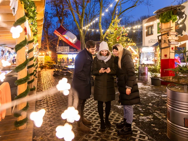 Wintermarkt bei Herr Kauzig - Weihnachten in Leipzig Drei Personen stehen auf dem Gelände des kauzigen Wintermarktes während im Hintergrund die festlich beleuchteten Verkaufsstände und der große Weihnachtsbaum zu sehen sind