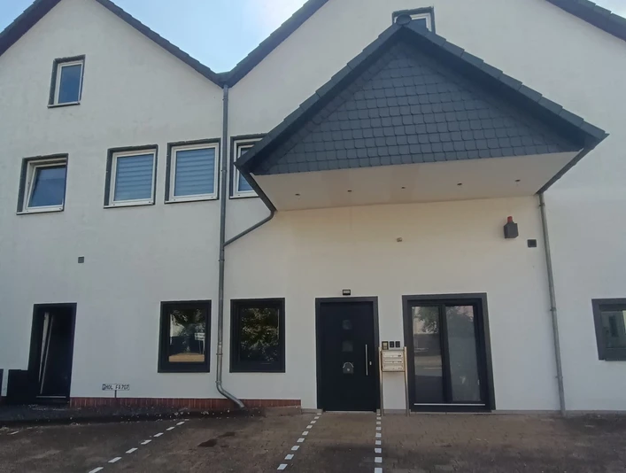 Frontansicht des Appartements asado Übderdachter Eingangsbereich eines modernen Gebäudes mit vielen Fenstern, Parkplätze vor dem Haus