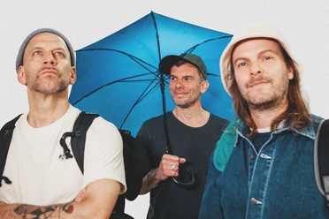 sportfreunde-stiller-c-paul-ambrusch-quer-cmyk.jpg Drei Männer mit Rucksäcken, zwei tragen Mützen, einer hält einen blauen Regenschirm und lächelt.