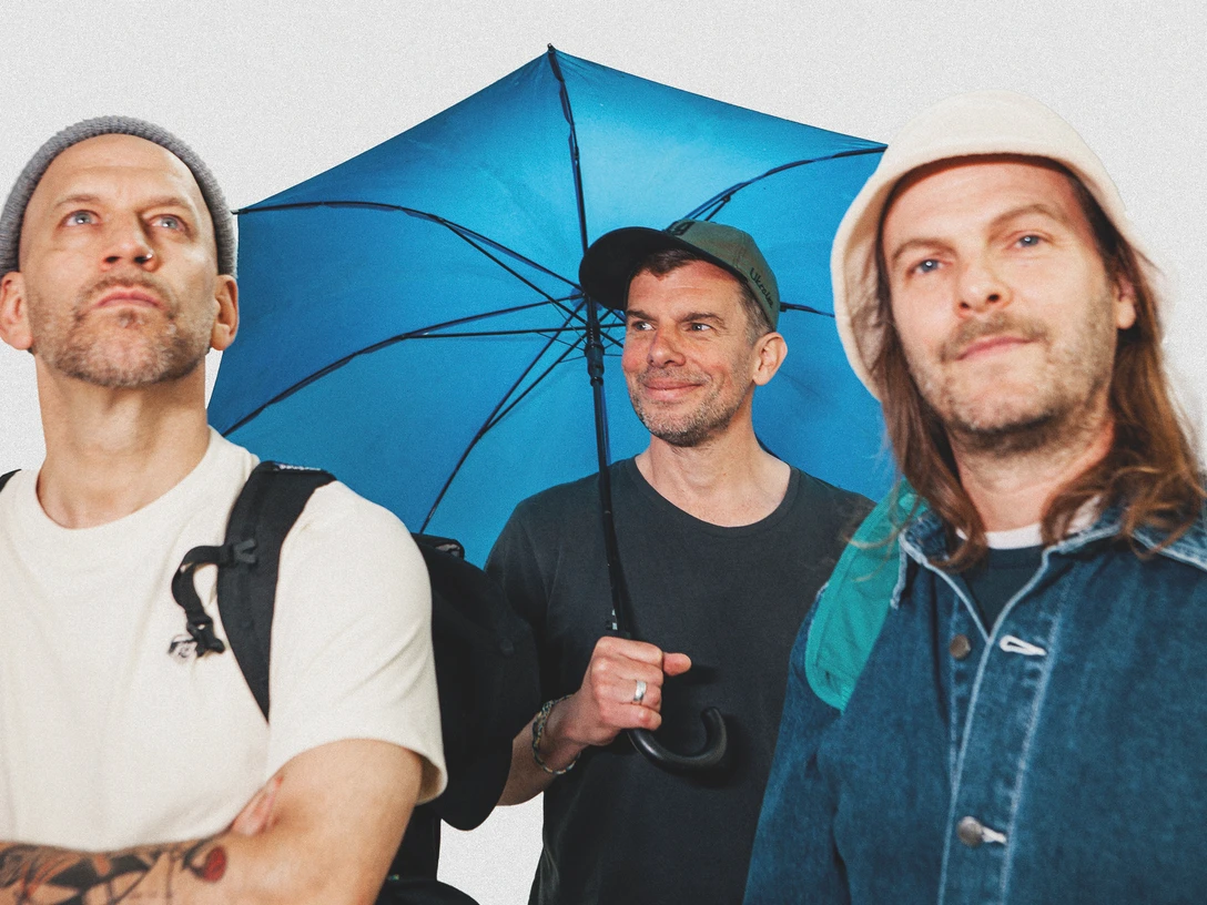 sportfreunde-stiller-c-paul-ambrusch-quer-cmyk.jpg Drei Männer mit Rucksäcken, zwei tragen Mützen, einer hält einen blauen Regenschirm und lächelt.