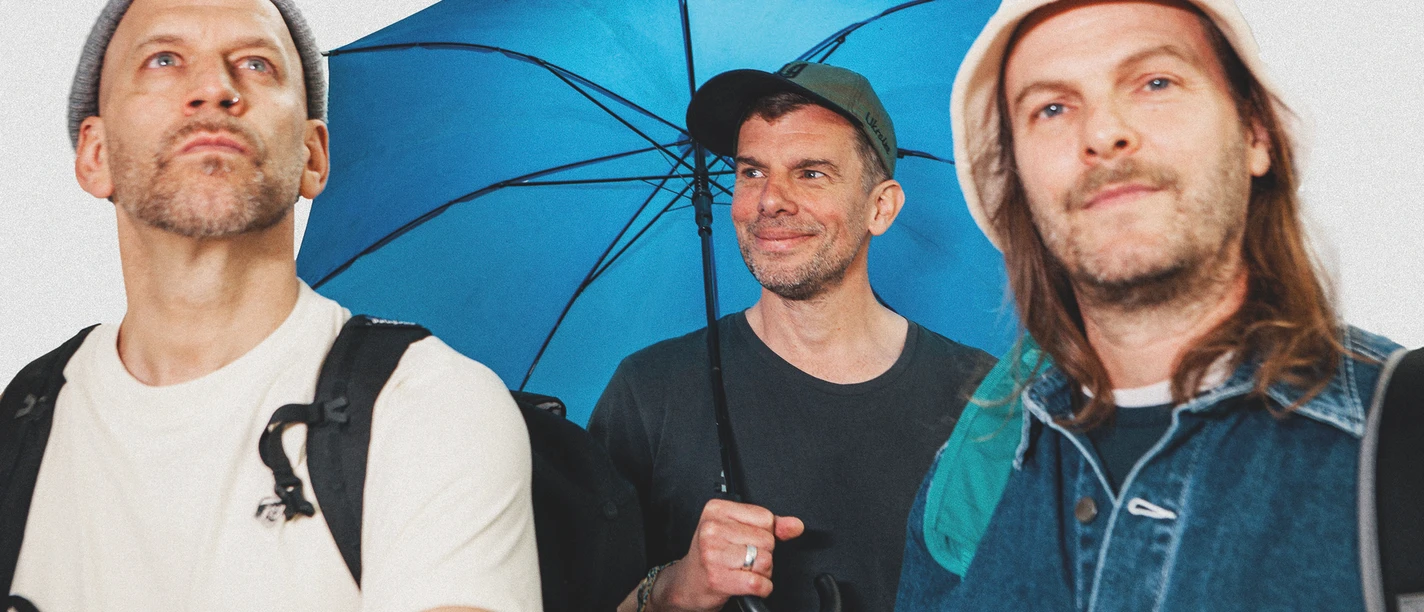 sportfreunde-stiller-c-paul-ambrusch-quer-cmyk.jpg Drei Männer mit Rucksäcken, zwei tragen Mützen, einer hält einen blauen Regenschirm und lächelt.