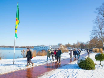 adventsspaziergang2 Menschen spazieren an einem verschneiten Seeufer mit Flagge und Ausflugsschiff im Wintersonnenschein.