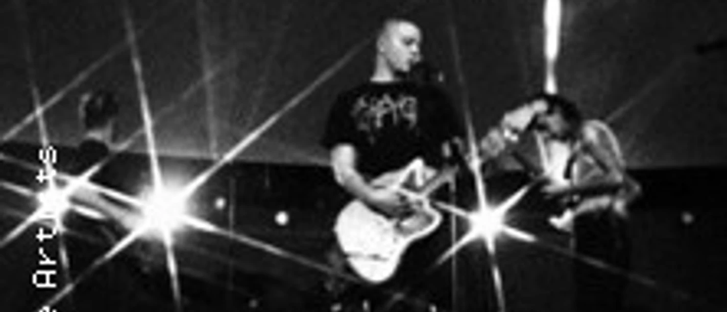 Drei Personen mit Gitarren bei einem Live-Konzert mit intensivem Bühnenlicht.