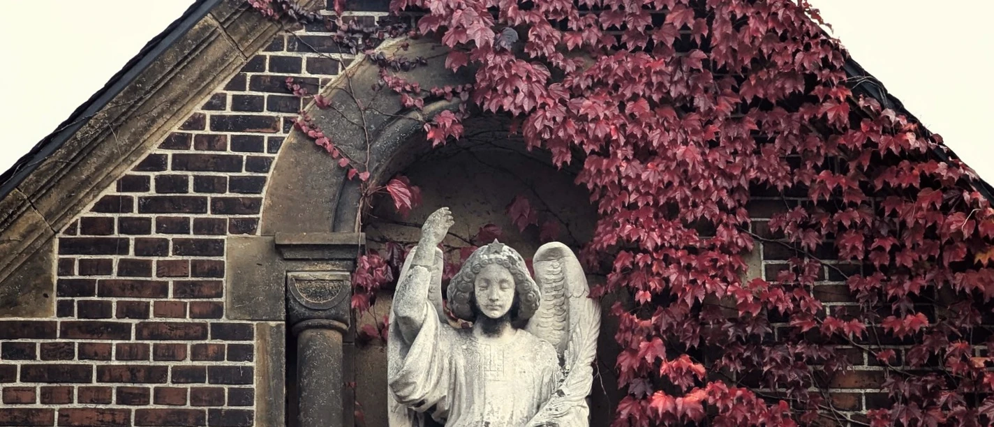 ©Bastian Ulrich - Nordfriedhof 1 Ein steinerner Engel wacht über eine mit roten Ranken bedeckte Ziegelwand unter einem Kreuz.