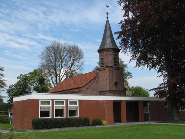 Kath. Kirche Augustfehn, Außenansicht von vorn, Foto Kath. Kirche.JPG Backsteinkirche mit kleinem Spitzturm und rotem Ziegeldach, umgeben von Rasen und Bäumen.