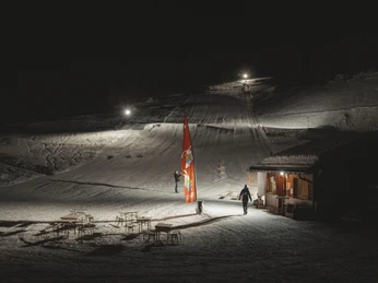 Atmospheric night skiing at the Fieschertal family ski lift Das Nachtskifahren Fieschertal zeigt die beleuchtete Talstation des Familienskilifts Blaetz mit Skifahrern und gemütlicher Hüttenatmosphäre in der Aletsch ArenaNight skiing Fieschertal shows the illuminated valley station of the Blaetz family ski lift with skiers and a cozy hut atmosphere in the Aletsch ArenaLe ski nocturne de Fieschertal montre la station inférieure éclairée du téléski familial Blaetz avec des skieurs et une ambiance chaleureuse dans un chalet de l'Aletsch Arena.