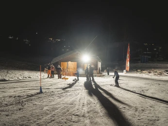 Téléski et lumière des chalets lors du ski nocturne à Fieschertal Das Nachtskifahren Fieschertal zeigt den beleuchteten Skilift Blaetz mit Skifahrern und Besuchern im warmen Licht der Aletsch Arena in der NachtNight skiing Fieschertal shows the illuminated Blaetz ski lift with skiers and visitors in the warm light of the Aletsch Arena at nightLe ski nocturne de Fieschertal montre le téléski Blaetz illuminé avec des skieurs et des visiteurs dans la lumière chaude de l'Aletsch Arena pendant la nuit.