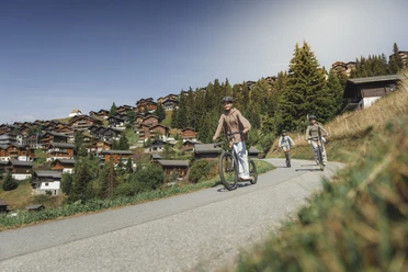 Trottinett-Abfahrt durch das autofreie Dorf Bettmeralp Beim Trottinett Aletsch Arena fahren drei Freunde auf sonnigen Wegen durch das autofreie Bergdorf Bettmeralp mit Blick auf die Walliser Alpen