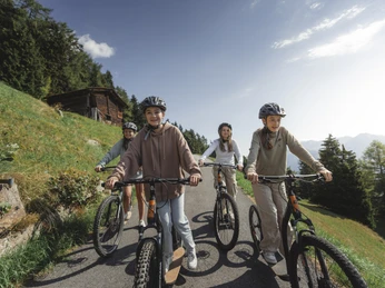 Des amis profitent d'une balade en trottinette dans l'Aletsch Arena Beim Trottinett Aletsch Arena fahren junge Freunde gemeinsam auf sonnigen Wegen unterhalb der Bettmeralp und geniessen den Blick auf die BergeOn the Aletsch Arena scooter, young friends ride together on sunny paths below Bettmeralp and enjoy the view of the mountainsLa trottinette Aletsch Arena permet à de jeunes amis de rouler ensemble sur des chemins ensoleillés en contrebas de Bettmeralp et d'admirer la vue sur les montagnes.