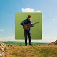 Ein Musiker spielt Gitarre auf einem Hügel vor blauem Himmel und grüner Landschaft.