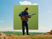Ein Musiker spielt Gitarre auf einem Hügel vor blauem Himmel und grüner Landschaft.