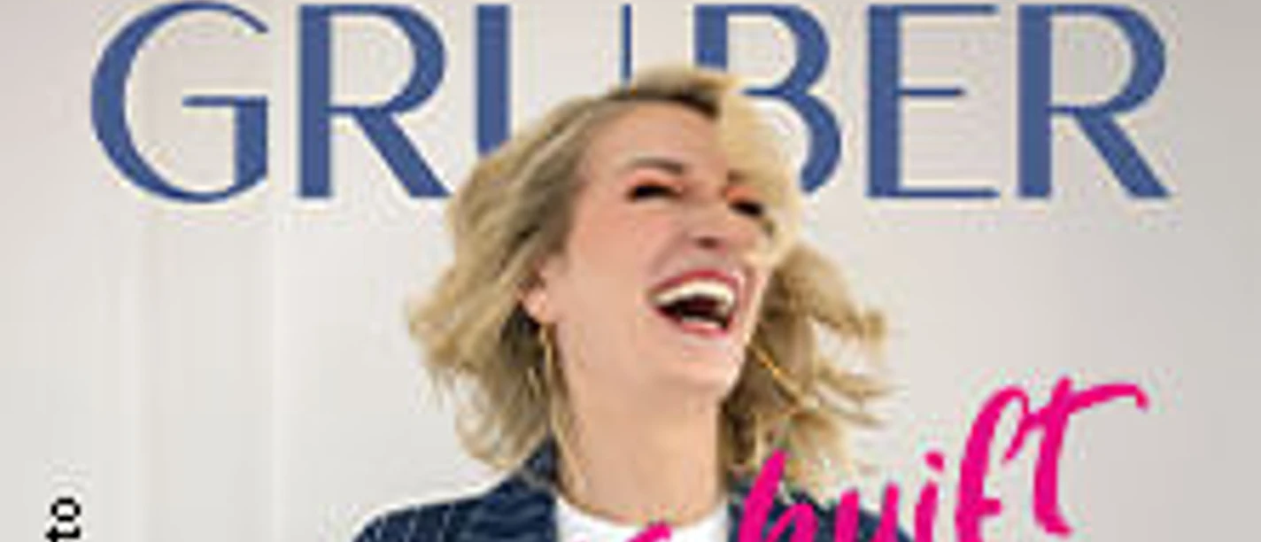 Monika Gruber lacht herzlich vor einer weißen Wand, neben ihr steht "es huift ja nix" in Rosa.