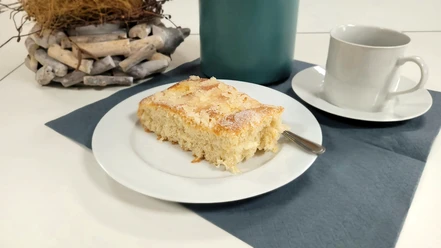 Kaffee und Kuchen.jpg Ein Stück Butterkuchen auf weißem Teller mit Kaffeetasse und blauem Untersetzer auf Tisch.