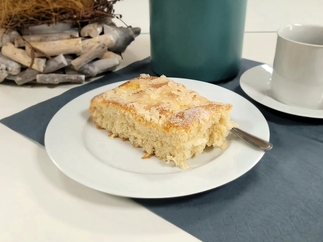 Kaffee und Kuchen.jpg Ein Stück Butterkuchen auf weißem Teller mit Kaffeetasse und blauem Untersetzer auf Tisch.