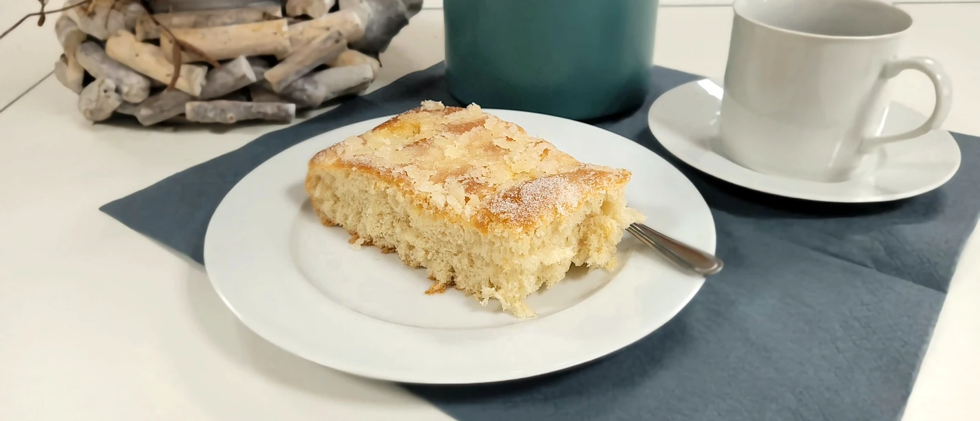 Kaffee und Kuchen.jpg Ein Stück Butterkuchen auf weißem Teller mit Kaffeetasse und blauem Untersetzer auf Tisch.