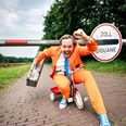 Patrick Nederkoorn (C) Nico Brons Mann im orangefarbenen Anzug auf kleinem Dreirad, lacht, hält Koffer, vor Zollschranke im Freien.