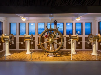 Steuerbrücke - Titanic: Eine Immersive Reise Nachbildung der Steuerbrücke mit Steuerrad und Geschwindigkeitsregler in der Ausstellung "Titanic: Eine Immersive Reise"Replica of the steering bridge with steering wheel and cruise control in the exhibition "Titanic: An Immersive Voyage"Replika kormidelního můstku s volantem a tempomatem na výstavě "Titanic: An Immersive Voyage" (Titanic: Plavba, která vás pohltí)Replika mostka sterowego z kierownicą i tempomatem na wystawie "Titanic: Wciągająca podróż"Replica van de stuurbrug met stuurwiel en cruisecontrol in de tentoonstelling "Titanic: An Immersive Voyage".Replica della plancia di comando con volante e cruise control nella mostra "Titanic: An Immersive Voyage"