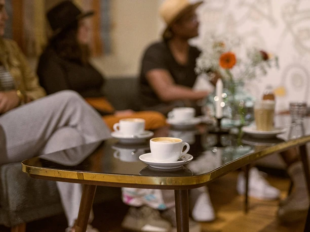 Das KAFF, Mülheim an der Ruhr Ein Couchtisch mit mehreren Tassen Kaffee und einer Gruppe junger Erwachsener im Hintergrund, die sich unterhalten.