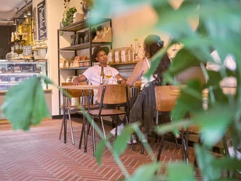 Ein Café mit modernem Interieur und einer entspannten Atmosphäre. Zwei Personen unterhalten sich an einem Tisch, umgeben von grünen Pflanzen und moderner Dekoration.