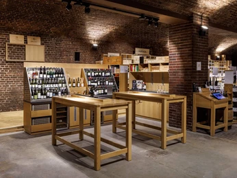 Kölner Weinkeller Kellerraum mit Weinen, Holzregalen und Verkostungstischen aus hellem Holz, umgeben von Backsteinwänden.Cellar room with wines, wooden shelves and tasting tables made of light-colored wood, surrounded by brick walls.