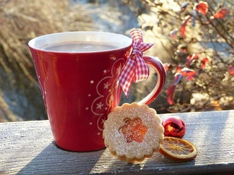 Rote Weihnachtstasse mit Schleife, Gebäck, Orangenscheibe und Kugel im winterlichen Sonnenlicht.