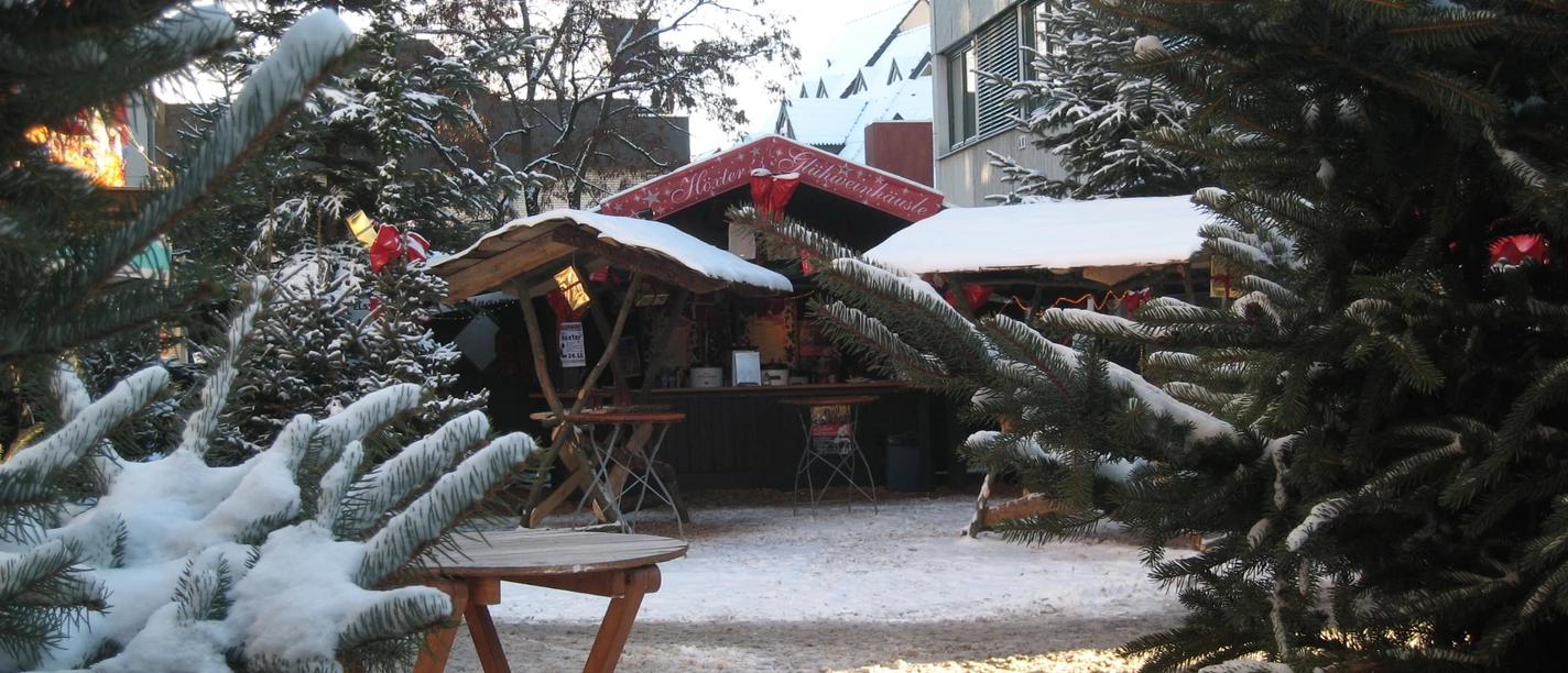 Verschneiter Weihnachtsmarkt mit Holzbuden, umgeben von Tannenbäumen, stimmungsvoll dekoriert.