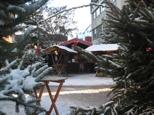 weihnachtsmarkt_winter_hubertusgrimm Verschneiter Weihnachtsmarkt mit Holzbuden, umgeben von Tannenbäumen, stimmungsvoll dekoriert.