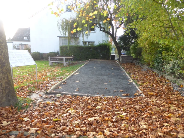Boulebahn im Herbst, angrenzend eine Rasenfläche, eine Informationstafel und eine Sitzgruppe. Im Hintergrund befindet sich ein Haus.