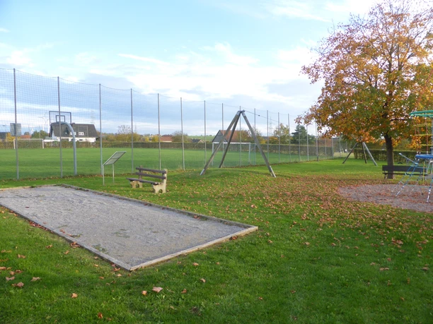 Die Boulebahn liegt umgeben von einer Rasenfläche direkt neben einem Spielplatz. Im Hintergrund ist ein Fußballplatz zu sehen.