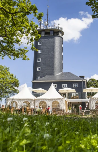 Hohe Bracht Ein markanter grauer Turm, umgeben von weißen Pavillons, vor einer Kulisse aus blühenden Wiesen.A striking gray tower, surrounded by white pavilions, against a backdrop of flowering meadows.Een opvallende grijze toren, omringd door witte paviljoens, tegen een achtergrond van bloeiende weiden.