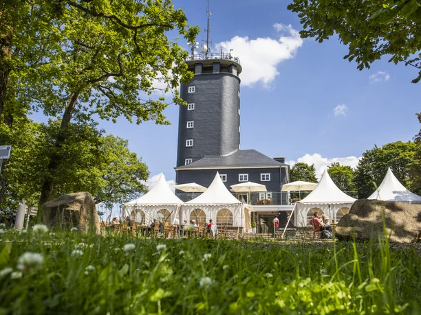 Hohe Bracht Ein markanter grauer Turm, umgeben von weißen Pavillons, vor einer Kulisse aus blühenden Wiesen.