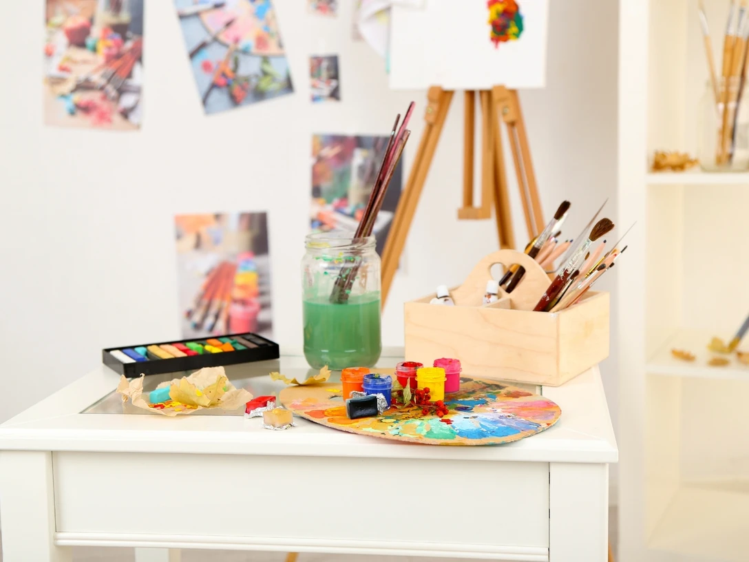 Kunstausstellung Im Bild sind diverse Utensilien zum Malen zu sehen (z.B. Farbpalette, Pinsel, Wasserglas, Aquarellfarben), die auf einem Tisch liegen. Im Hintergrund sieht man eine Staffelei und Bilder an der Wand hängend.