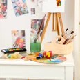 Kunstausstellung Im Bild sind diverse Utensilien zum Malen zu sehen (z.B. Farbpalette, Pinsel, Wasserglas, Aquarellfarben), die auf einem Tisch liegen. Im Hintergrund sieht man eine Staffelei und Bilder an der Wand hängend.