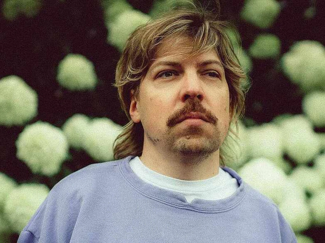 philipp-poisel-c-sophie-seybold-cmyk.png Person mit blondem Haar und Schnurrbart blickt ernst in die Ferne, im Hintergrund weiße Blüten.