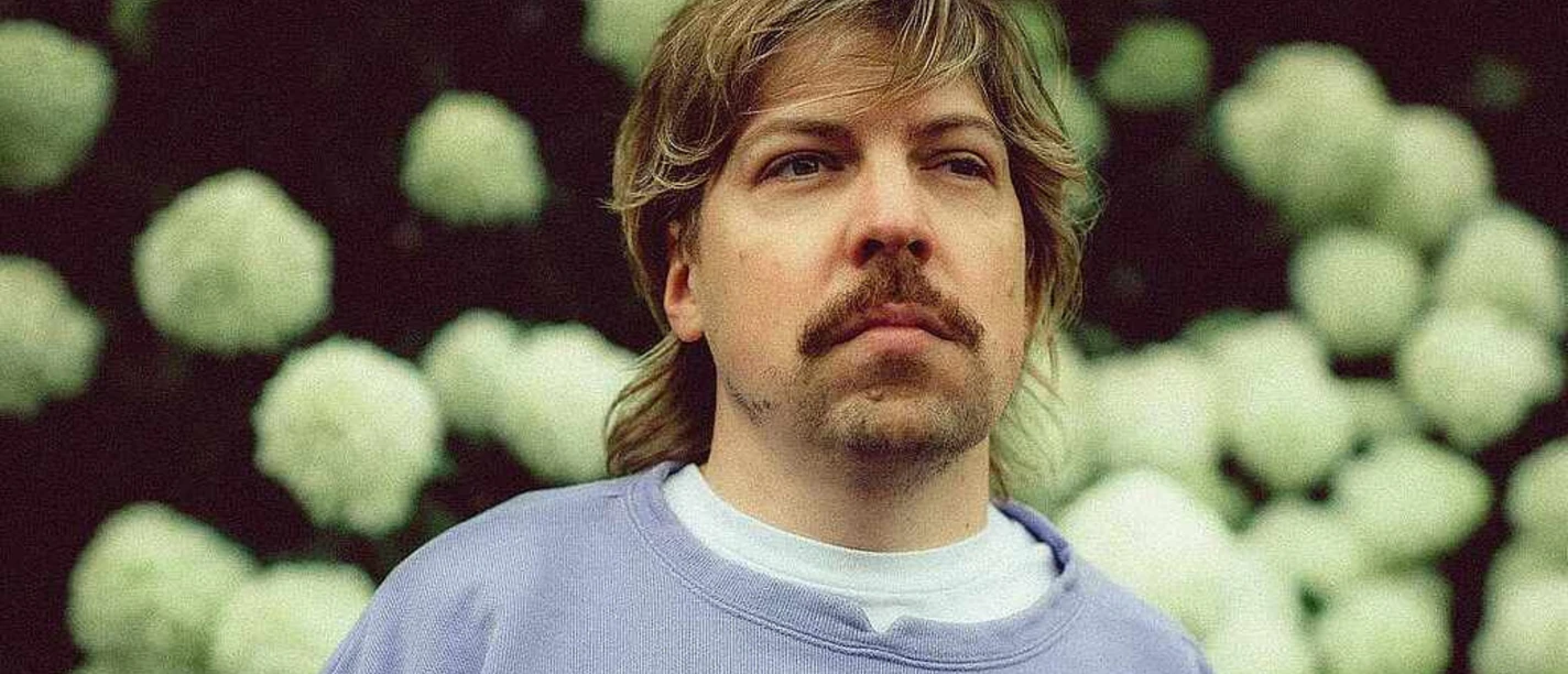 philipp-poisel-c-sophie-seybold-cmyk.png Person mit blondem Haar und Schnurrbart blickt ernst in die Ferne, im Hintergrund weiße Blüten.