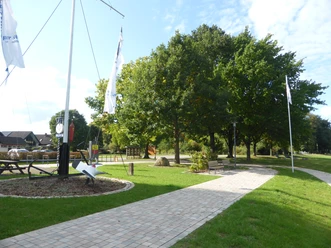 Gepflasterter Weg an der Weserpromenade in Wehrden mit Sitzmöglichkeiten, Fahnenmast, Schiffsanker und Bäumen.