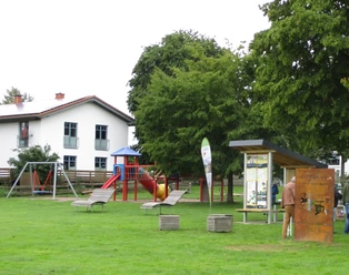 Rast-Info-Punkt an der Weserpromenade Würgassen Eine Rasenfläche mit Bäumen und Sitzwürfeln, einer Stele und einem überdachten Rast-Info-Punkt, im Hintergrund ein Spielplatz und ein Haus.