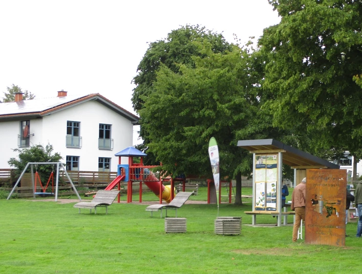 Rast-Info-Punkt an der Weserpromenade Würgassen Eine Rasenfläche mit Bäumen und Sitzwürfeln, einer Stele und einem überdachten Rast-Info-Punkt, im Hintergrund ein Spielplatz und ein Haus.