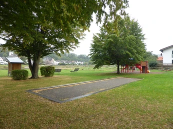 Eine Boulebahn auf einer Rasenfläche unter großen Bäumen, im Hintergrund ein überdachter Rast-Info-Punkt , ein Spielplatz und ein Haus.