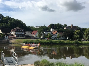 Eine Personenfähre überquert die Weser, im Vordergrund die Anlegestelle und im Hintergrund der Ort Herstelle.