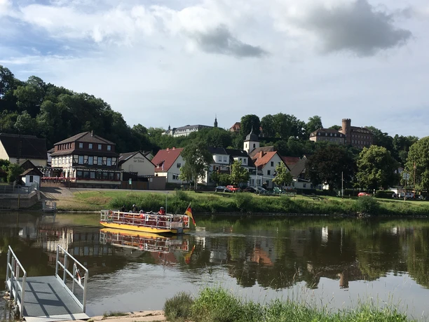 Eine Personenfähre überquert die Weser, im Vordergrund die Anlegestelle und im Hintergrund der Ort Herstelle.