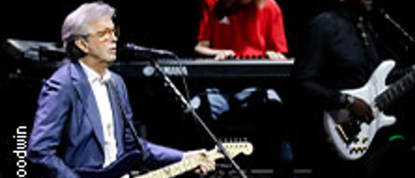 Eric Clapton spielt Gitarre auf einer Bühne mit Keyboarder und Bassist im Hintergrund.