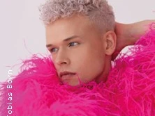 Eine Person mit hellblonden Locken trägt ein pinkfarbenes, federndes Outfit und blickt seitwärts.