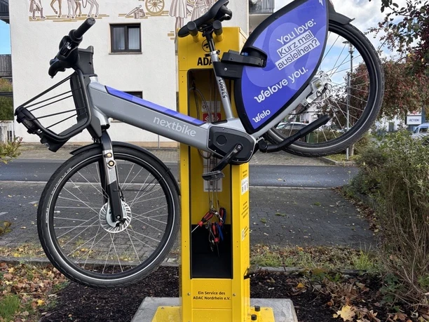 Radservice-Station Ruppichteroth An einer gelben Radservicestation hängt ein blaues Leihfahrrad der Marke Nextbike. Die Station steht an einem gepflasterten Gehweg vor einem Gebäude.