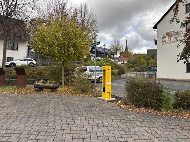 Radservice-Station Ruppichteroth Gelbe Radservice-Station im urbanen Umfeld, Bäume daneben, Gebäude im Hintergrund.