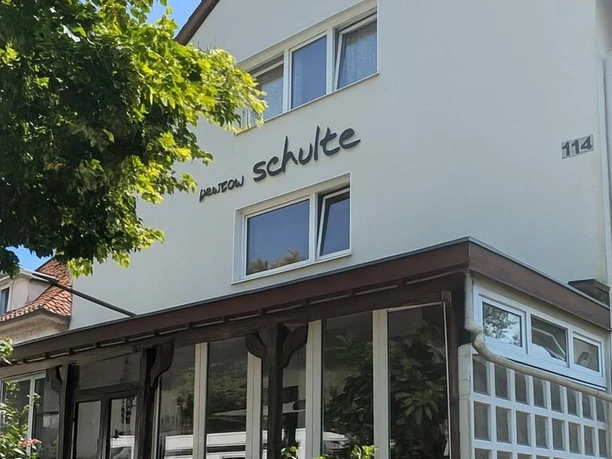 Ein Gebäude mit einem begrünten Vorgarten und großen Fenstern. Der Name "Pension Schulte" ist an der Fassade angebracht.