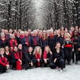 Rastede Gospel Choir.png Große fröhliche Chorgruppe in Schwarz und Rot posiert im verschneiten Wald mit kahlen Bäumen.