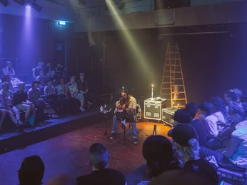 Bonanza festival Eine Person sitzt auf einem Stuhl in einem beleuchteten Raum voller Besucherinnen und Besucher. Im Hintergrund sind Leitern und Beleuchtungstechnik zu sehen.A person sits on a chair in an illuminated room full of visitors. Ladders and lighting equipment can be seen in the background.