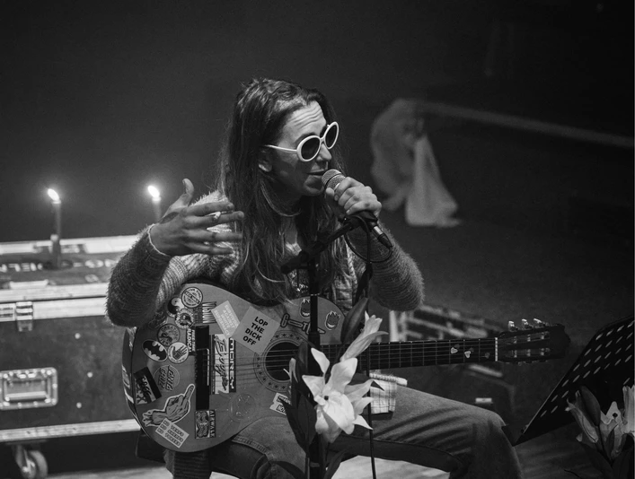 Bonanza festival Eine Person sitzt auf einem Stuhl und spielt Gitarre, während sie singt. Sie trägt eine Sonnenbrille und ist umgeben von Kerzenlicht.A person sits on a chair and plays the guitar while singing. She is wearing sunglasses and is surrounded by candlelight.