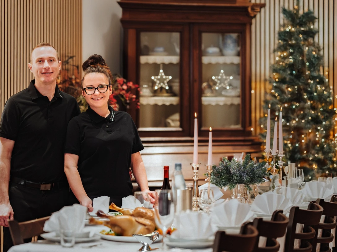 Spargelhof Winkelmann Zwei Mitarbeitende in schwarzer Kleidung stehen vor festlich gedecktem Tisch mit Kerzen und Tannenschmuck.